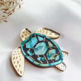 A handcrafted sea turtle brooch with a blue and turquoise shell and cream-colored legs and head, placed on white satin fabric with some dried plant stems to the side, showcases an artistic design.
