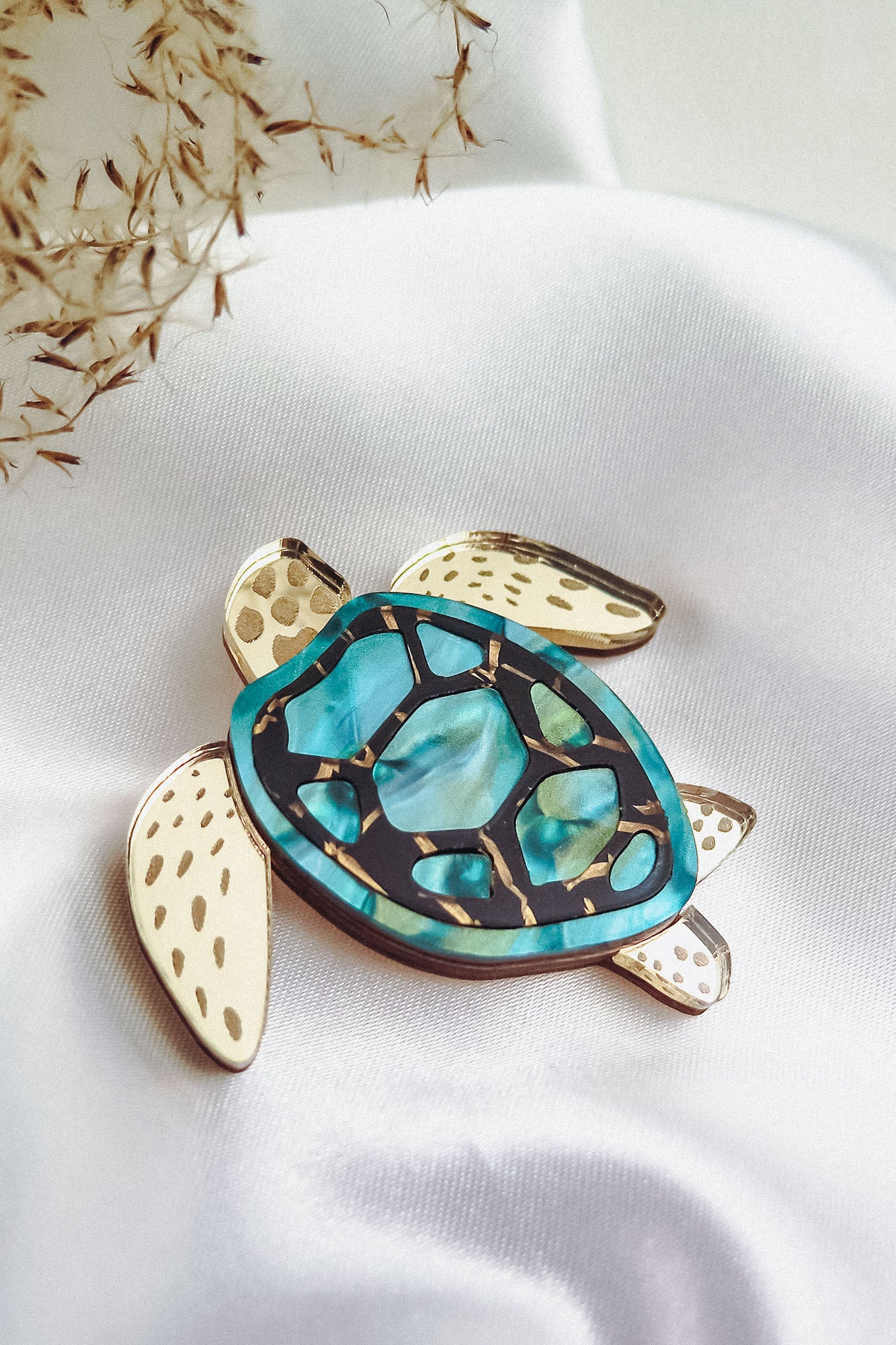 A handcrafted sea turtle brooch with a blue and turquoise shell and cream-colored legs and head, placed on white satin fabric with some dried plant stems to the side, showcases an artistic design.