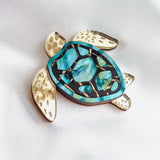 A handcrafted sea turtle brooch with gold-toned limbs and a turquoise, blue, and black artistic design on its shell, displayed on a white fabric background.