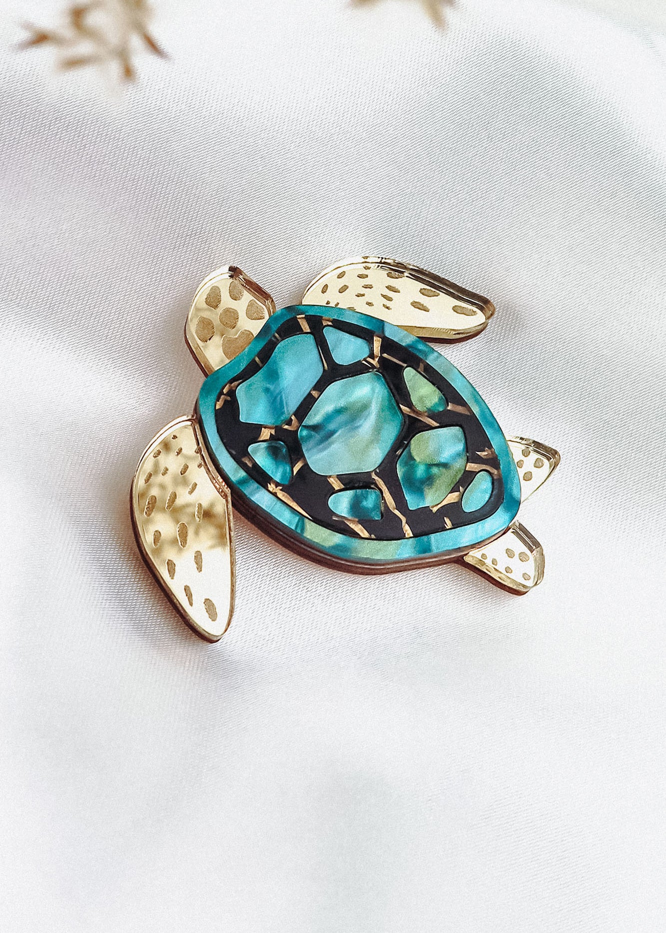 A handcrafted sea turtle brooch with gold-toned limbs and a turquoise, blue, and black artistic design on its shell, displayed on a white fabric background.