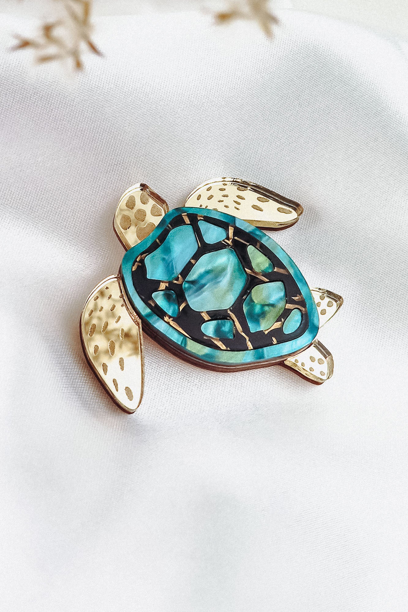 A handcrafted sea turtle brooch with gold-toned limbs and a turquoise, blue, and black artistic design on its shell, displayed on a white fabric background.