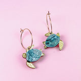 Handcrafted Little Turtle Hoop Earrings featuring gold hoops and blue and gold sea turtle charms, displayed on a pink background.