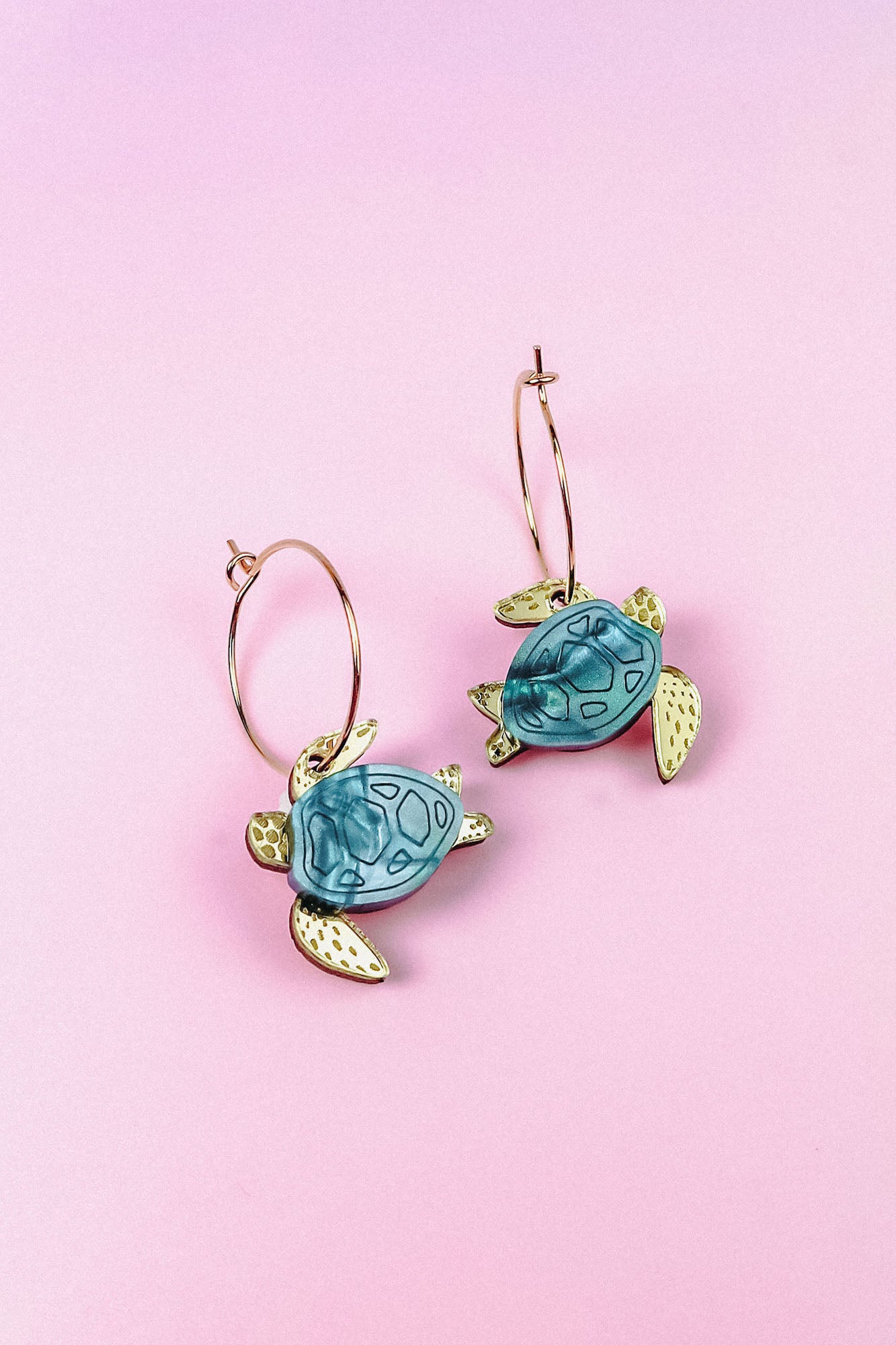 Handcrafted Little Turtle Hoop Earrings featuring gold hoops and blue and gold sea turtle charms, displayed on a pink background.