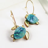 Gold hoops featuring blue and gold handcrafted turtle-shaped charms with detailed shells and flippers, the Little Turtle Hoop Earrings are displayed on a white background.