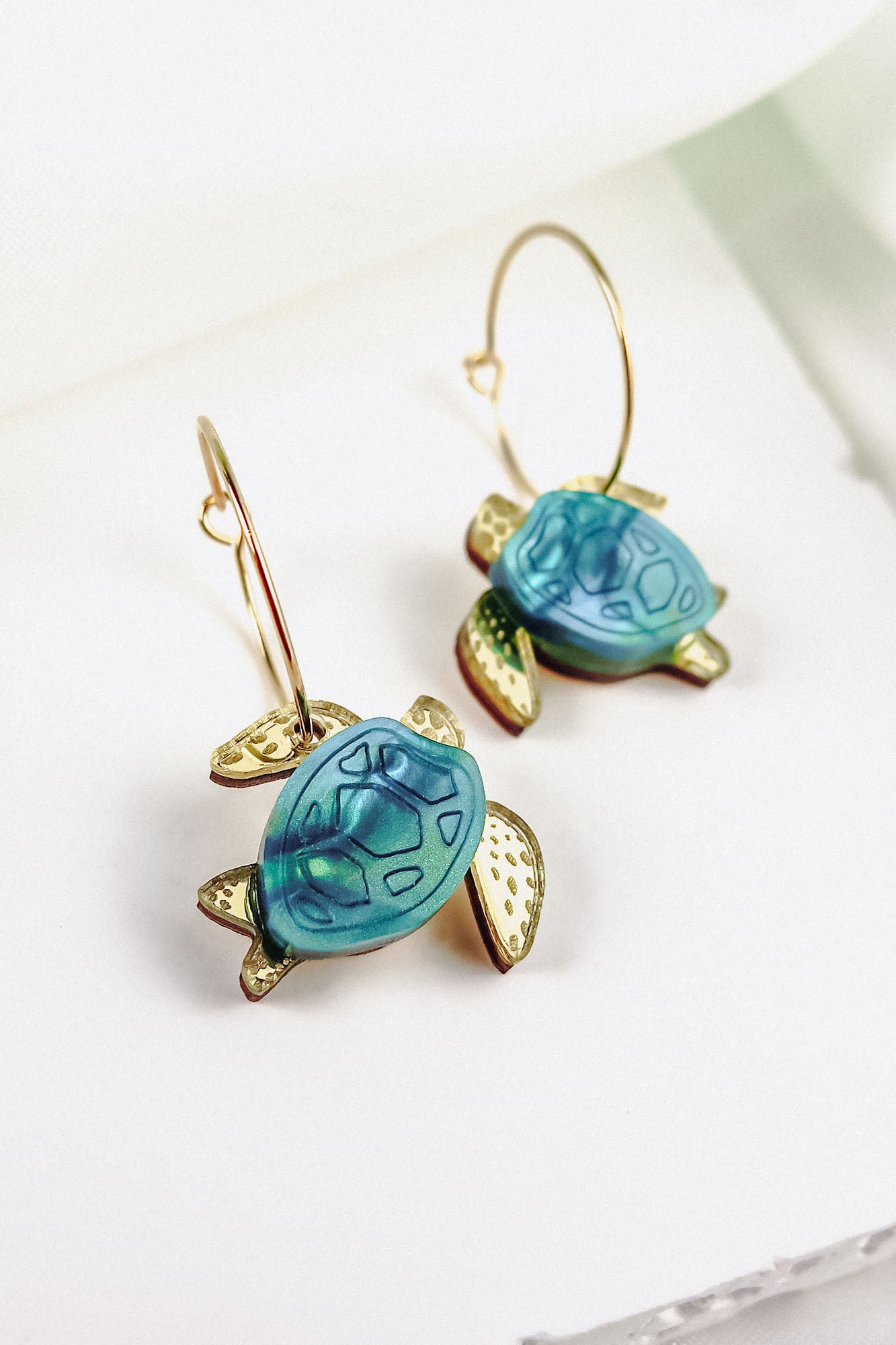 Gold hoops featuring blue and gold handcrafted turtle-shaped charms with detailed shells and flippers, the Little Turtle Hoop Earrings are displayed on a white background.