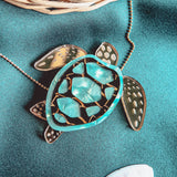 A close-up of a handcrafted Sea Turtle Necklace pendant with a turquoise and gold shell, lying on green fabric next to a white seashell and a wicker basket.