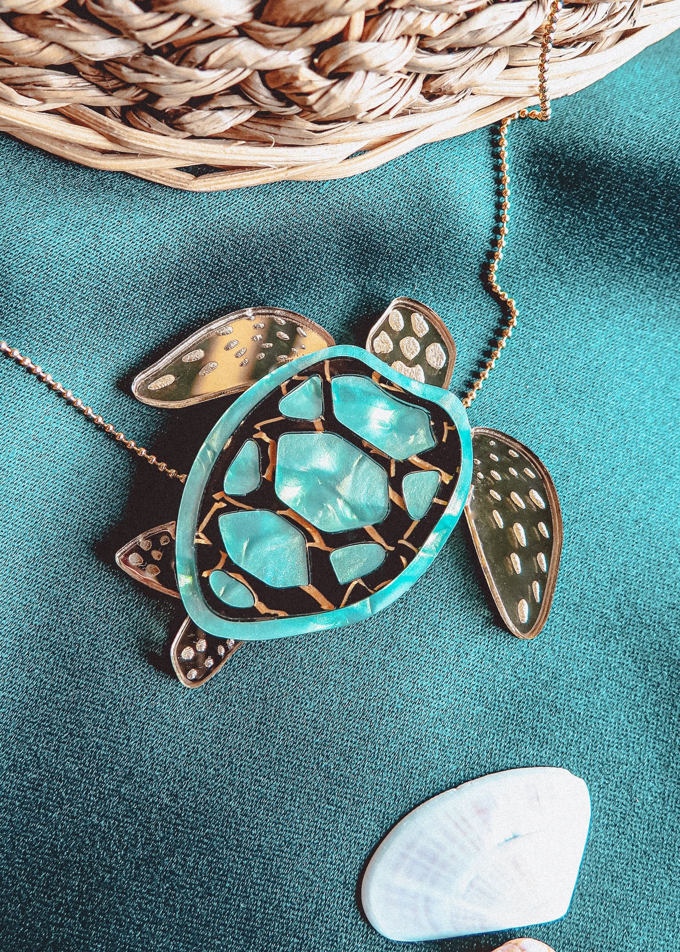 A close-up of a handcrafted Sea Turtle Necklace pendant with a turquoise and gold shell, lying on green fabric next to a white seashell and a wicker basket.