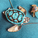 A close-up of a shiny handcrafted Sea Turtle Necklace with a turquoise shell and gold accents, lying on a teal fabric surface next to a woven basket and another small accessory.