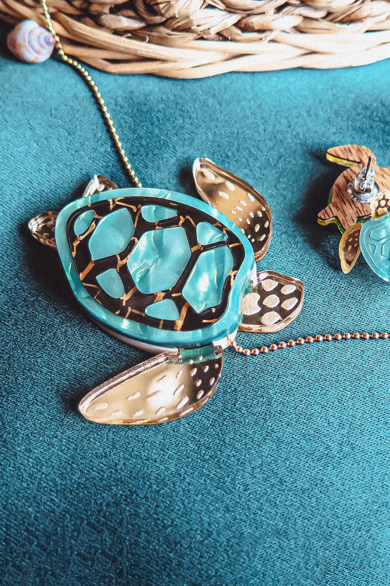 A close-up of a shiny handcrafted Sea Turtle Necklace with a turquoise shell and gold accents, lying on a teal fabric surface next to a woven basket and another small accessory.
