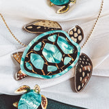 A handcrafted gold necklace featuring a turquoise and gold sea turtle pendant, accompanied by two matching small turtle charms, beautifully displayed on white fabric.