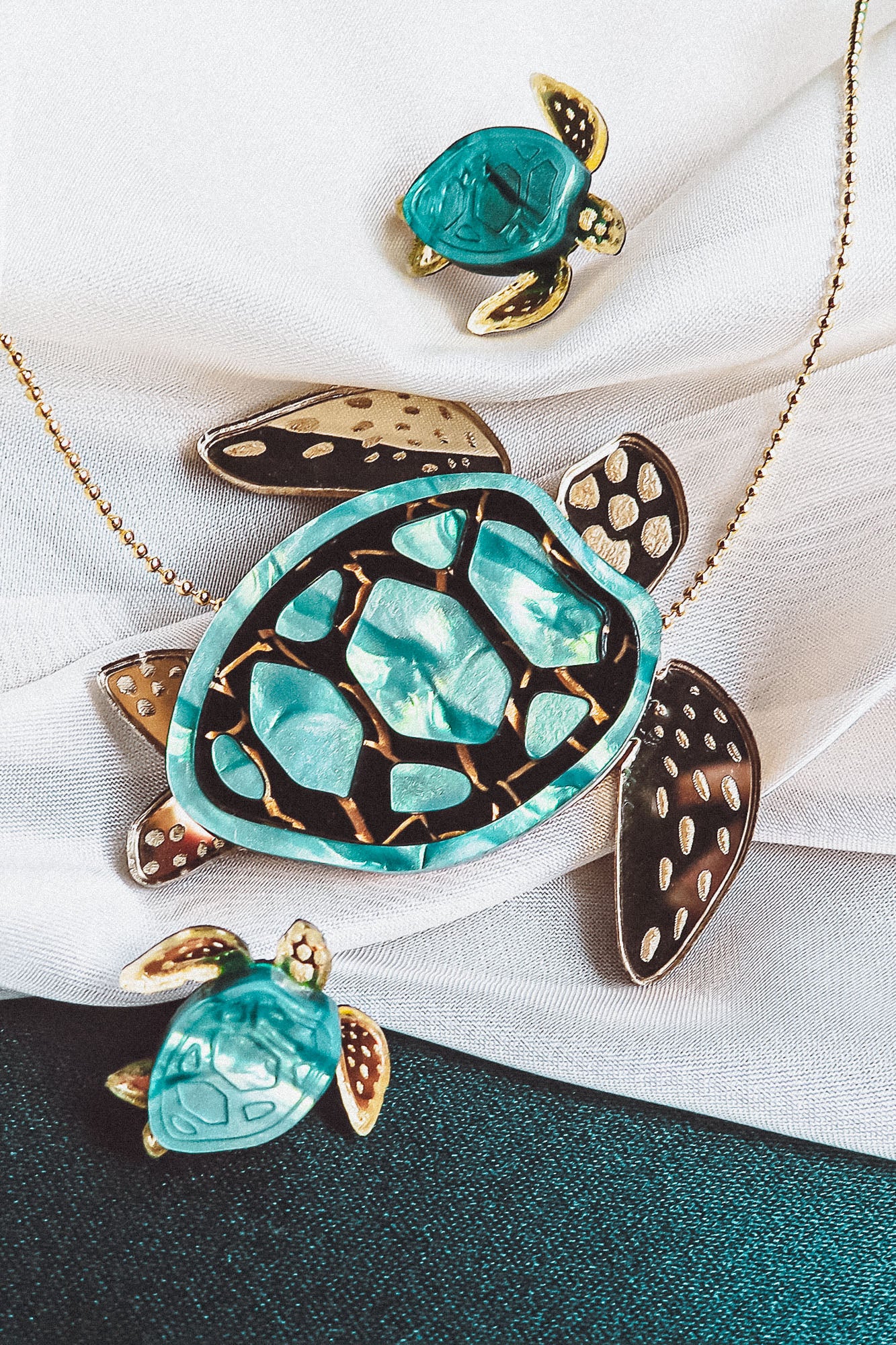 A handcrafted gold necklace featuring a turquoise and gold sea turtle pendant, accompanied by two matching small turtle charms, beautifully displayed on white fabric.