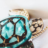 Close-up of a handcrafted sea turtle necklace featuring a turquoise shell with gold accents, resting on a white textured fabric background.