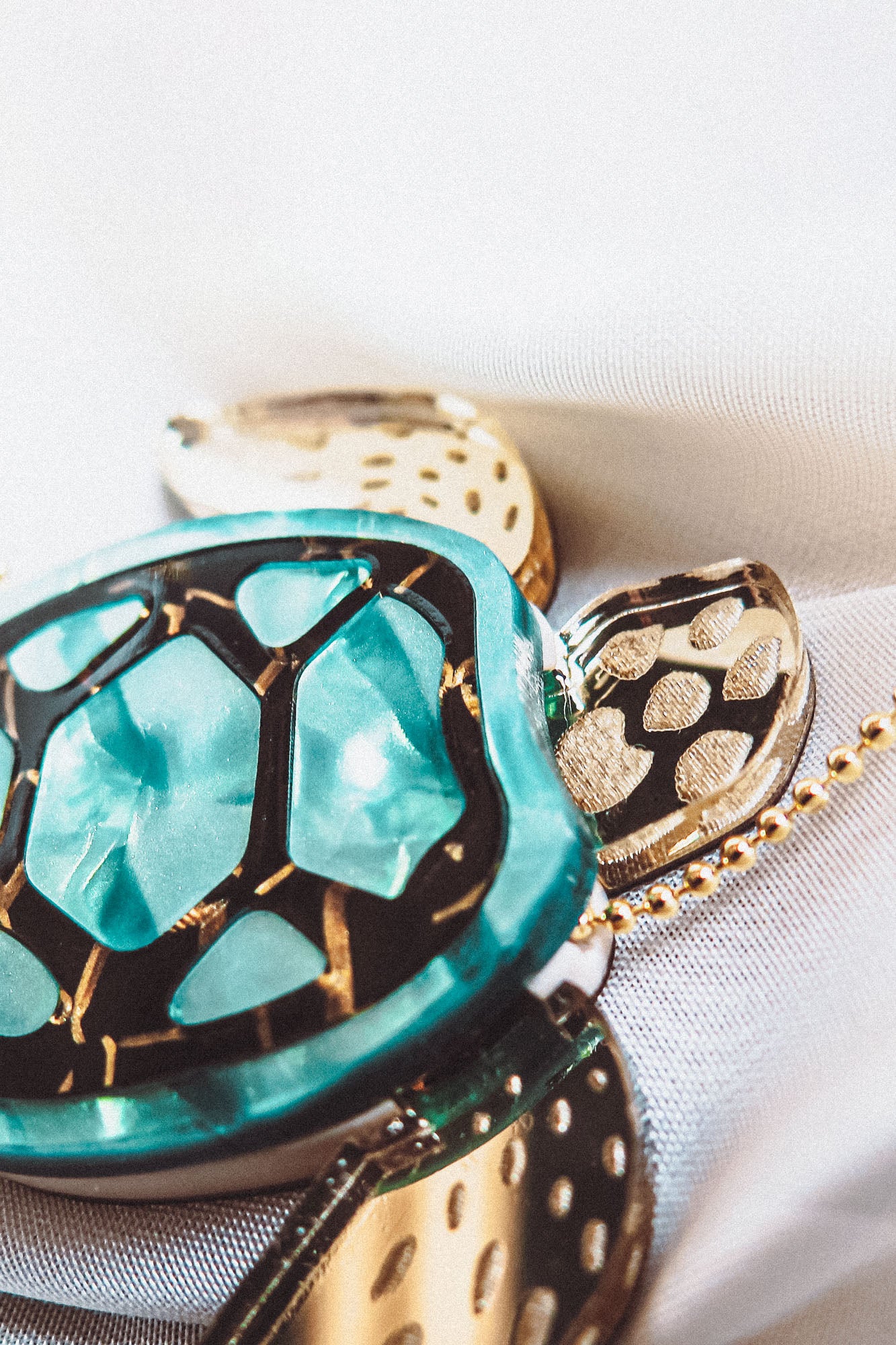 Close-up of a handcrafted sea turtle necklace featuring a turquoise shell with gold accents, resting on a white textured fabric background.