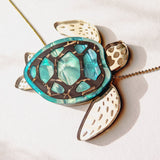 A close-up of a handcrafted Sea Turtle Necklace with a turquoise shell and white flippers, lying on a light surface. The turtle has intricate shell details and a gold chain.