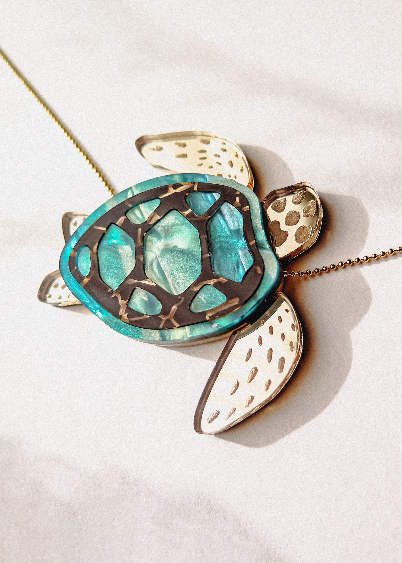 A close-up of a handcrafted Sea Turtle Necklace with a turquoise shell and white flippers, lying on a light surface. The turtle has intricate shell details and a gold chain.