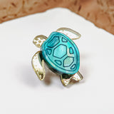 A gold and turquoise sea turtle-shaped ring, handcrafted with mirror-acrylic glass, rests on a white surface against a blurred brown background.
