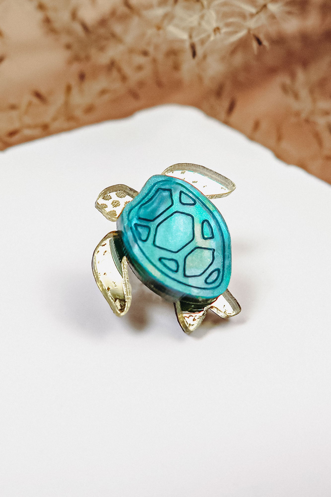 A gold and turquoise sea turtle-shaped ring, handcrafted with mirror-acrylic glass, rests on a white surface against a blurred brown background.