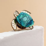 A handcrafted sea turtle pin with a blue-green shell and gold-toned, textured flippers is perched on the edge of a white textured surface against a beige background.