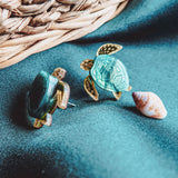 Two Sea Turtle Studs with green shells rest on a teal fabric beside a small seashell, evoking an ocean-inspired, summery vibe. A woven basket is visible in the background.