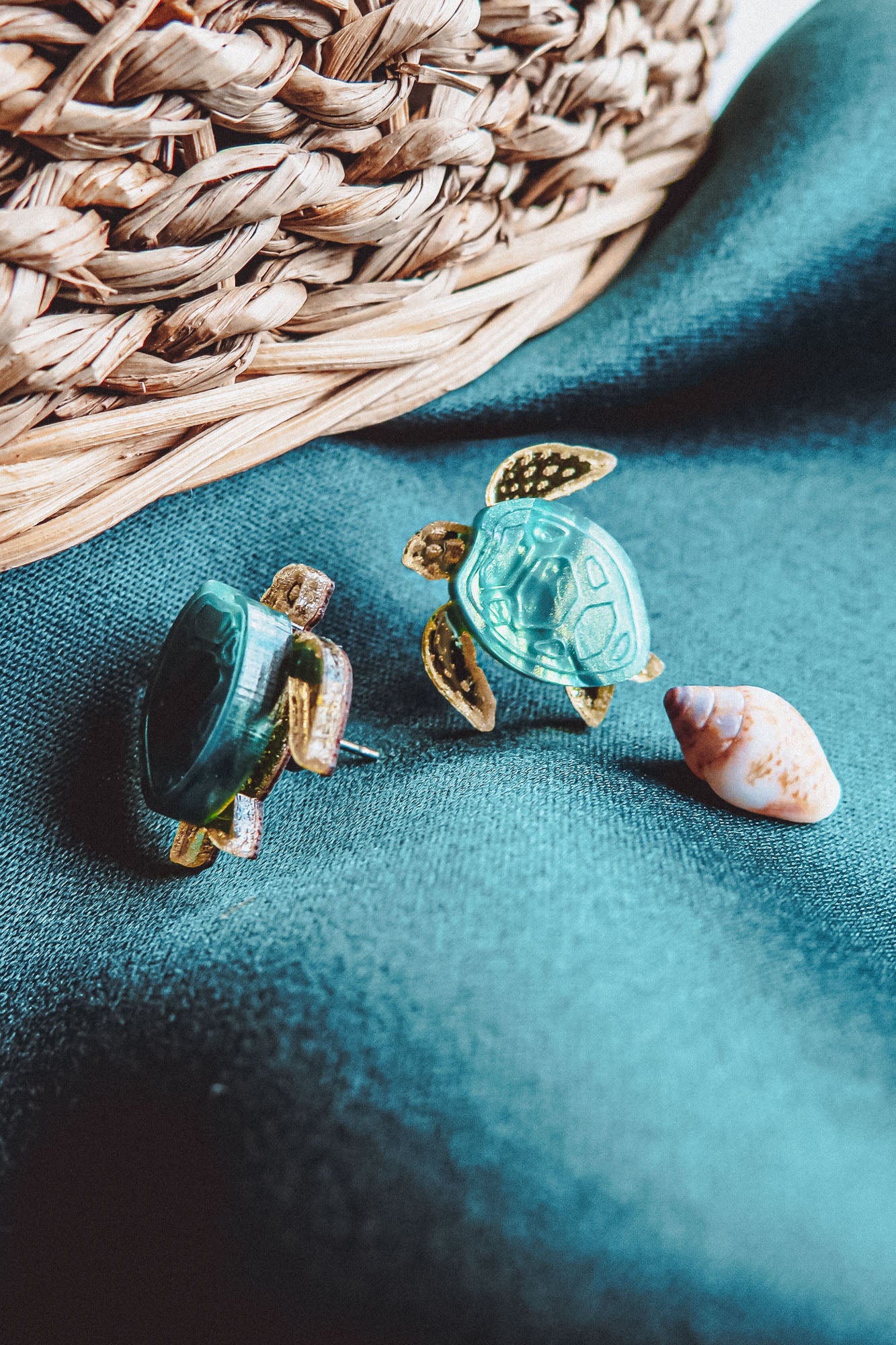 Two Sea Turtle Studs with green shells rest on a teal fabric beside a small seashell, evoking an ocean-inspired, summery vibe. A woven basket is visible in the background.