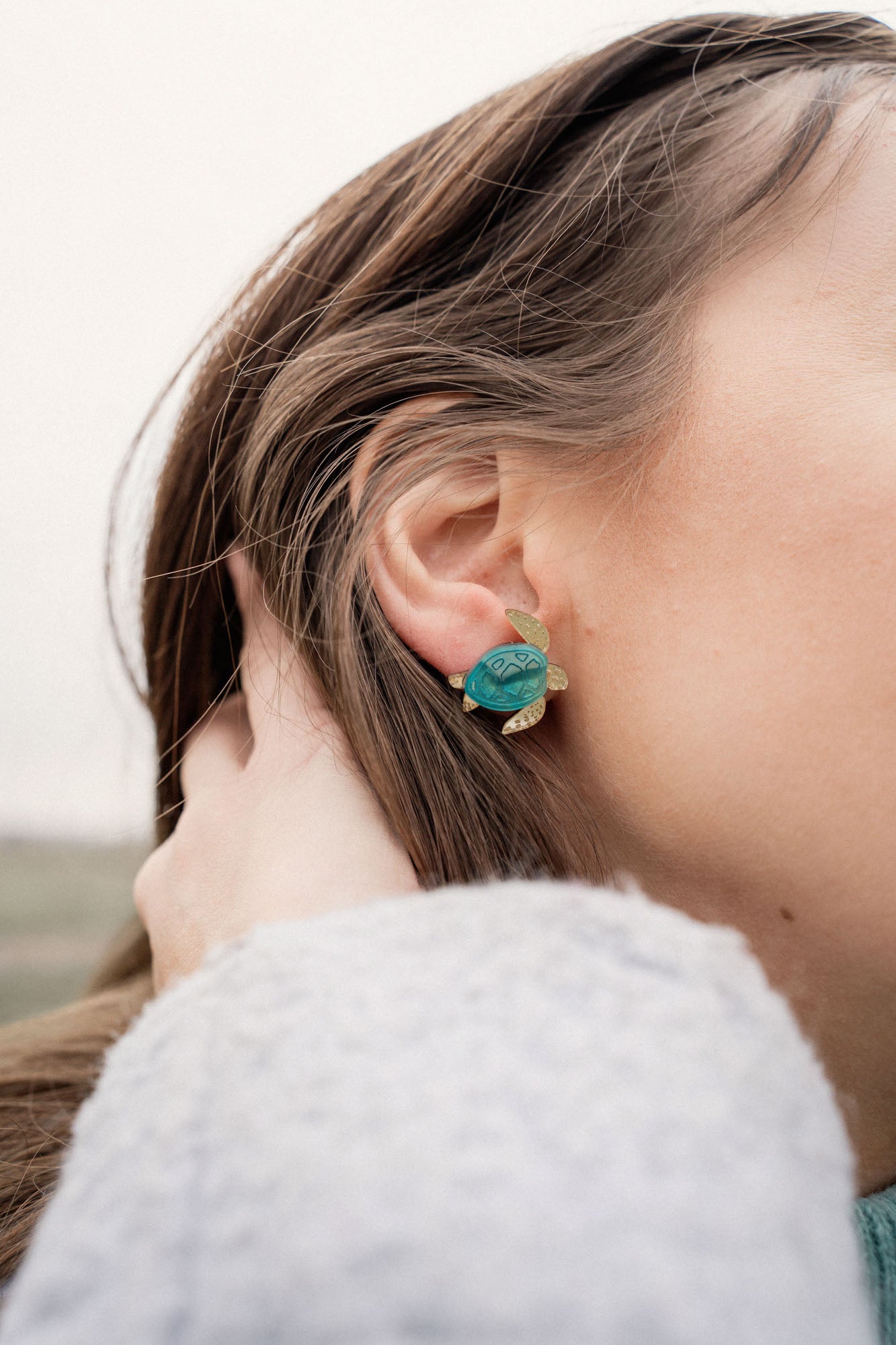 A close-up of a persons ear wearing gold and turquoise Sea Turtle Studs, with their light brown hair partially visible, captures an ocean-inspired, summery vibe.