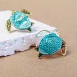 Two gold and turquoise Sea Turtle Studs rest on a piece of textured white material against a beige background, bringing an ocean-inspired, summery vibe to your accessories.