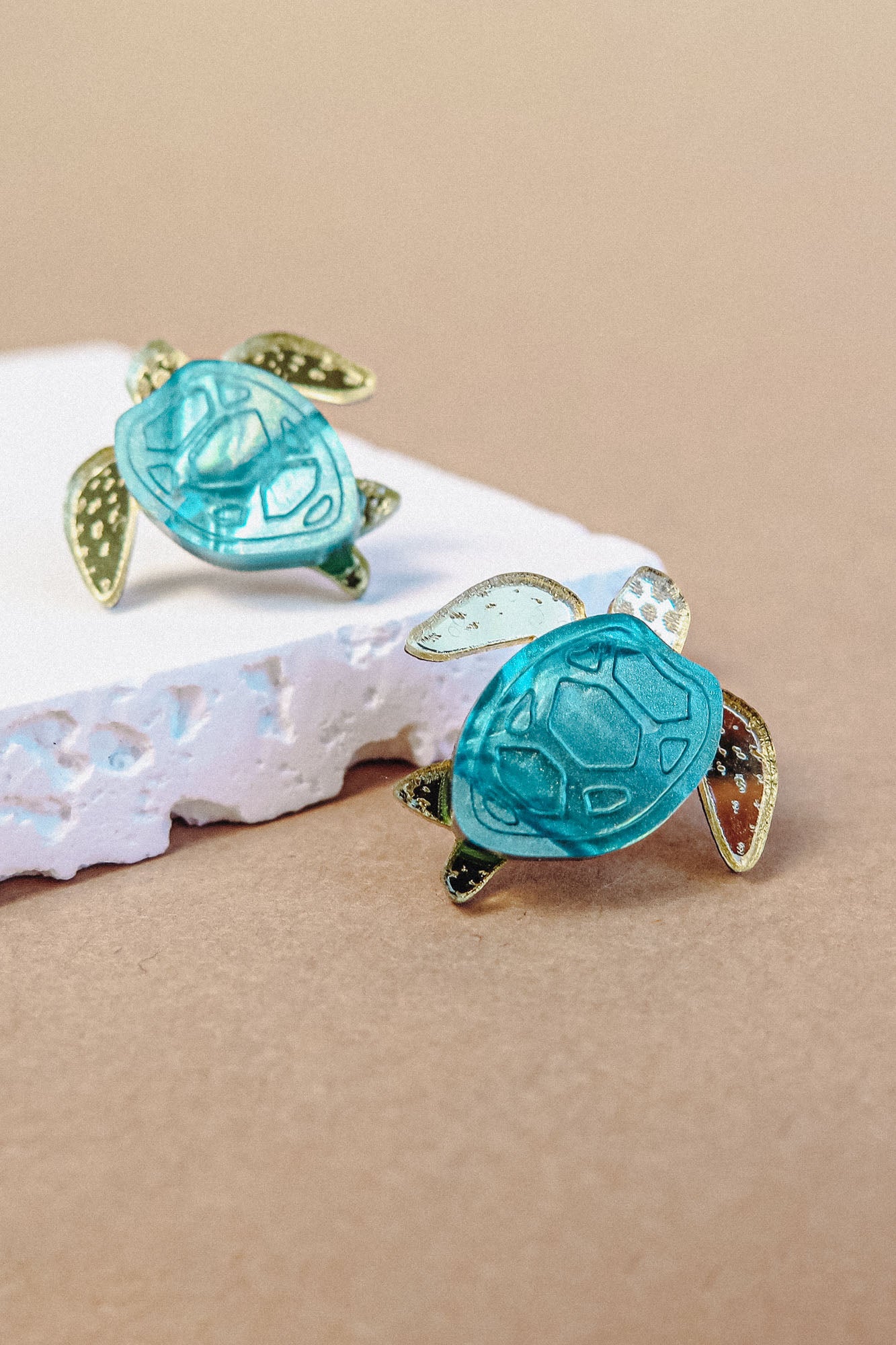 Two gold and turquoise Sea Turtle Studs rest on a piece of textured white material against a beige background, bringing an ocean-inspired, summery vibe to your accessories.