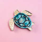 A handcrafted sea turtle brooch featuring an artistic design with a blue and teal shell and beige, patterned limbs, placed on a light pink background.