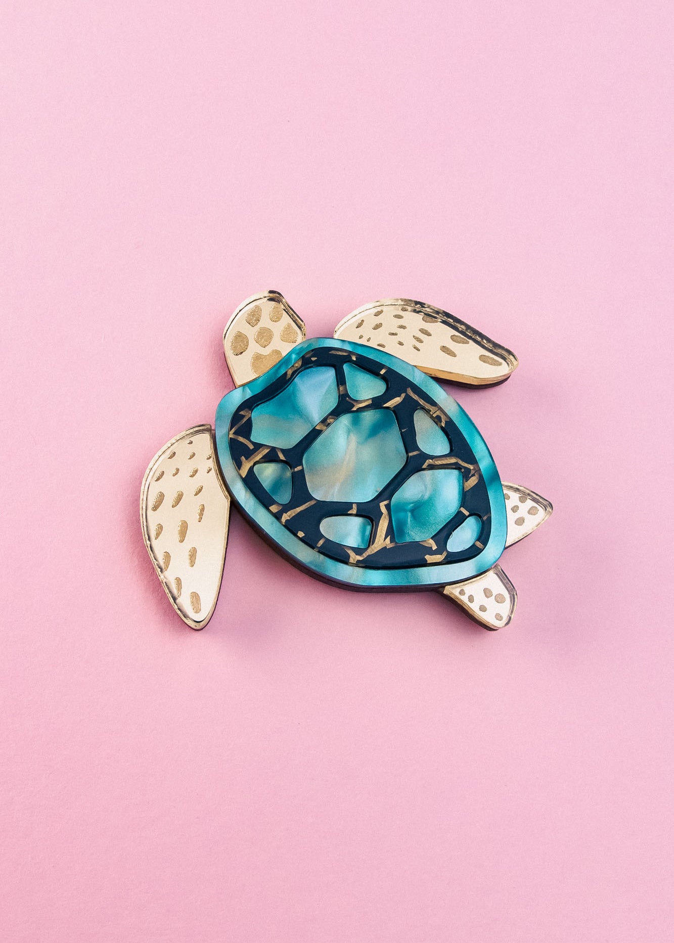 A handcrafted sea turtle brooch featuring an artistic design with a blue and teal shell and beige, patterned limbs, placed on a light pink background.