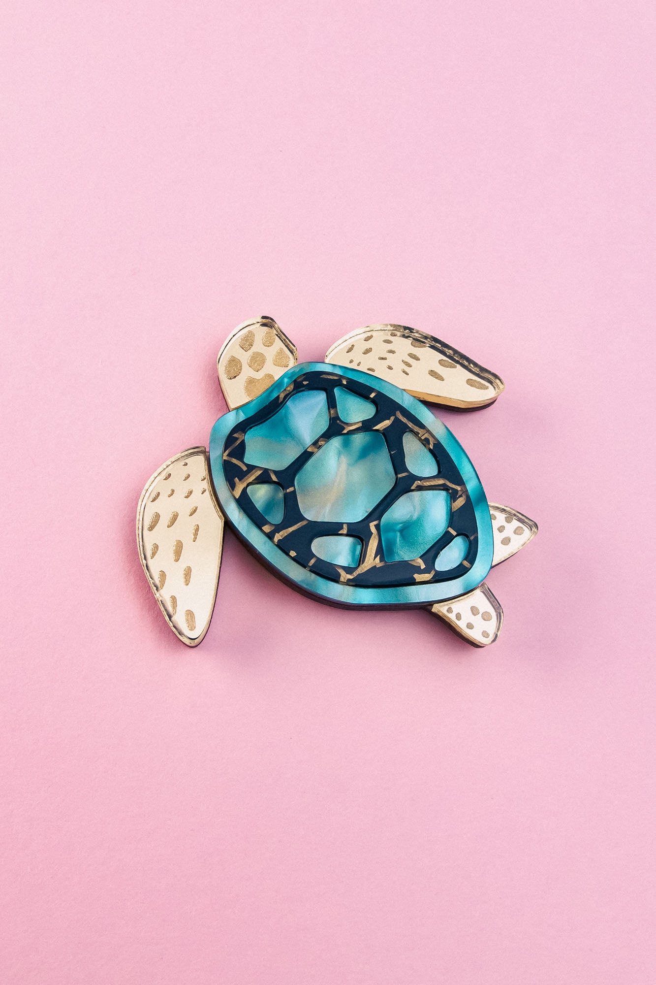 A handcrafted sea turtle brooch featuring an artistic design with a blue and teal shell and beige, patterned limbs, placed on a light pink background.