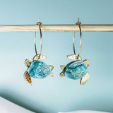 Two handcrafted gold hoops with hanging blue and gold turtle-shaped charms are displayed on a wooden rod against a light blue background, complemented by white fabric at the bottom. Discover the charm of our Little Turtle Hoop Earrings.