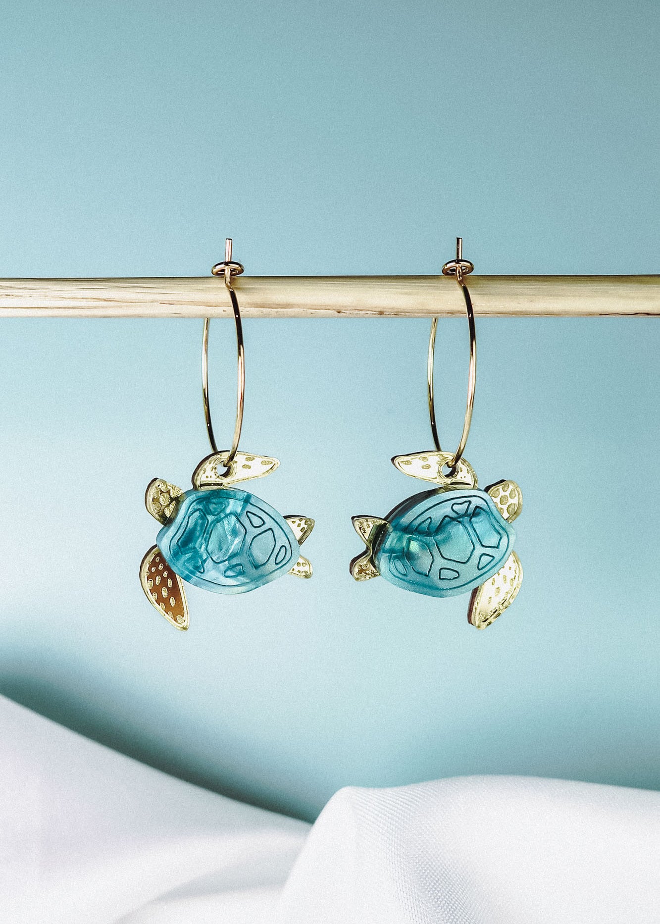 Two handcrafted gold hoops with hanging blue and gold turtle-shaped charms are displayed on a wooden rod against a light blue background, complemented by white fabric at the bottom. Discover the charm of our Little Turtle Hoop Earrings.
