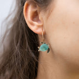 Close-up of a womans ear wearing Little Turtle Hoop Earrings—handcrafted gold hoops adorned with a turtle charm featuring a turquoise shell and gold flippers. The softly blurred background adds an elegant touch.