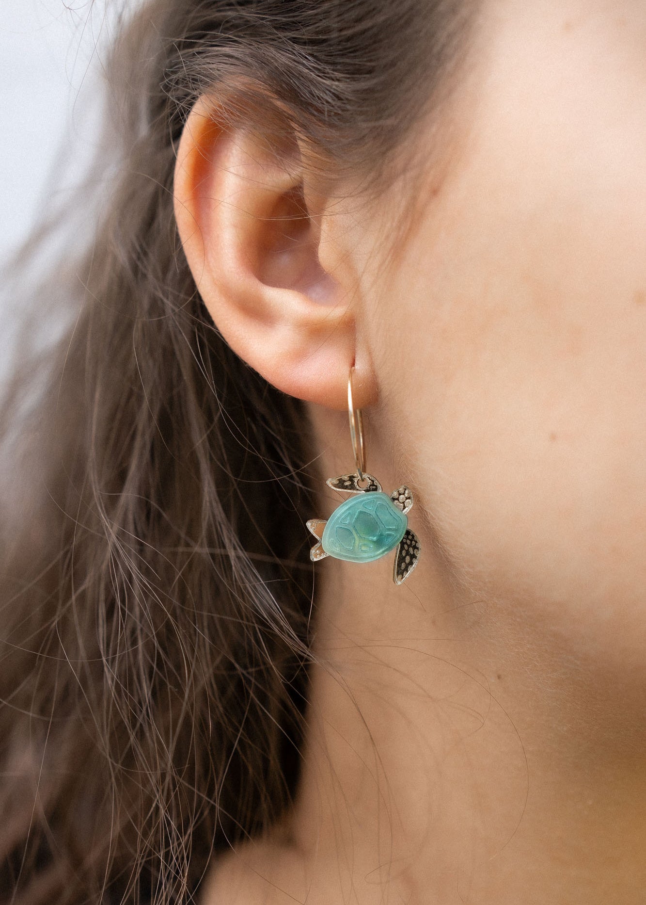 Close-up of a womans ear wearing Little Turtle Hoop Earrings—handcrafted gold hoops adorned with a turtle charm featuring a turquoise shell and gold flippers. The softly blurred background adds an elegant touch.