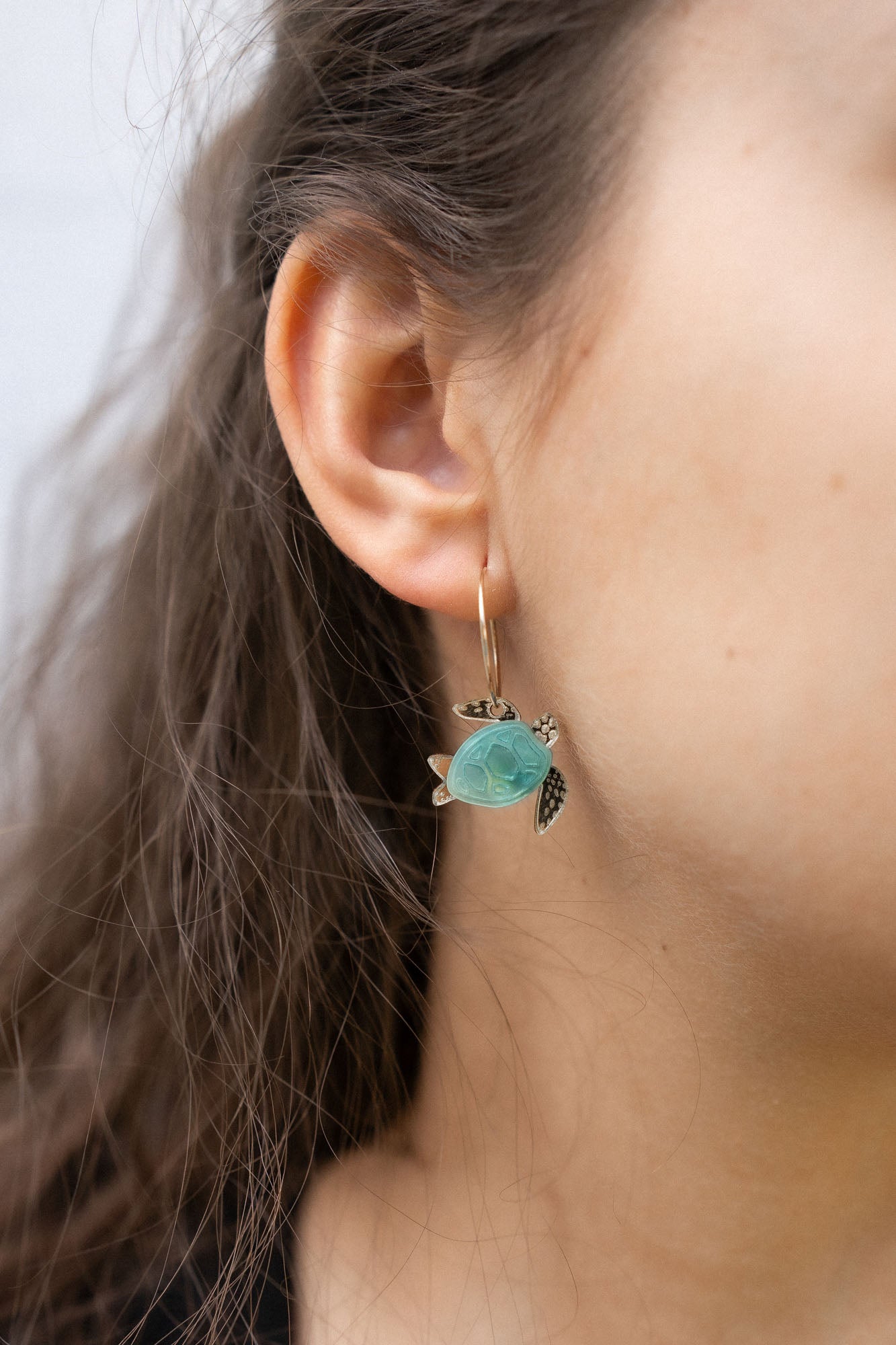 Close-up of a womans ear wearing Little Turtle Hoop Earrings—handcrafted gold hoops adorned with a turtle charm featuring a turquoise shell and gold flippers. The softly blurred background adds an elegant touch.