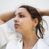 A woman with dark hair pulled back holds her hands behind her head, wearing a white blouse and handcrafted Little Turtle Hoop Earrings with green gemstones, looking thoughtfully into the distance against a plain light background.