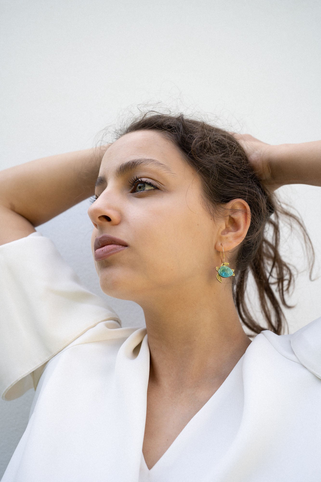 A woman with dark hair pulled back holds her hands behind her head, wearing a white blouse and handcrafted Little Turtle Hoop Earrings with green gemstones, looking thoughtfully into the distance against a plain light background.