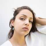 A young woman with light eyes and wavy brown hair poses against a plain white background, wearing a white top and handcrafted Little Turtle Hoop Earrings, with one hand touching her hair.