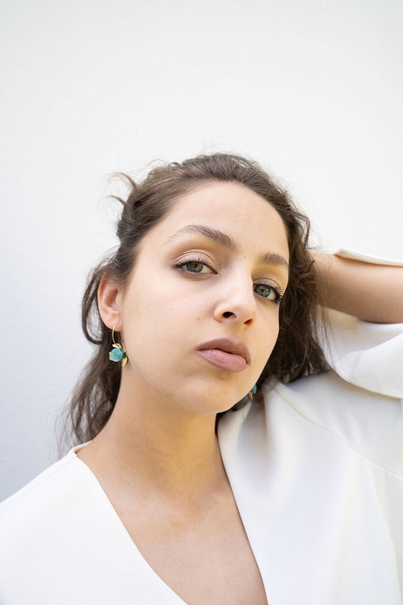 A young woman with light eyes and wavy brown hair poses against a plain white background, wearing a white top and handcrafted Little Turtle Hoop Earrings, with one hand touching her hair.