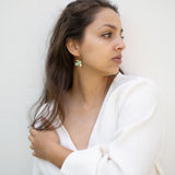 A woman with long brown hair wearing a white top and handcrafted Little Turtle Hoop Earrings looks to her right, standing against a plain white wall and gently holding her hair with one hand.