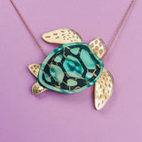 A handcrafted Sea Turtle Necklace with a pendant featuring a green and black shell, gold-colored flippers, and hints of turquoise, displayed against a purple background.