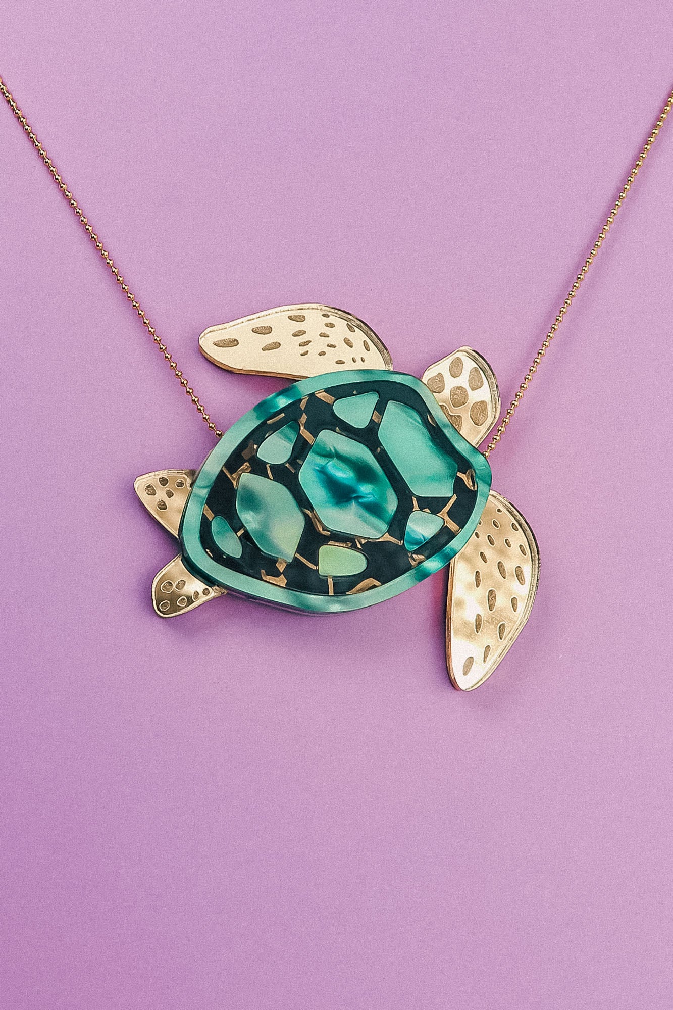 A handcrafted Sea Turtle Necklace with a pendant featuring a green and black shell, gold-colored flippers, and hints of turquoise, displayed against a purple background.