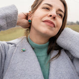 A woman in a light blue coat and teal sweater stands outdoors, smiling gently with her hands behind her head. She wears matching turquoise earrings and a handcrafted sea turtle pin made of mirror-acrylic glass, with a grassy field in the background.