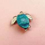 A small sea turtle pin, handcrafted with gold-colored flippers and a blue mirror-acrylic glass shell, rests on a light pink background.
