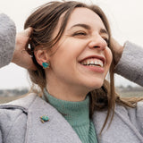 A woman with straight, light brown hair and light skin wears a light pink top and turquoise Sea Turtle Studs, standing in front of a mauve curtain, gazing slightly to the left for a summery vibe.