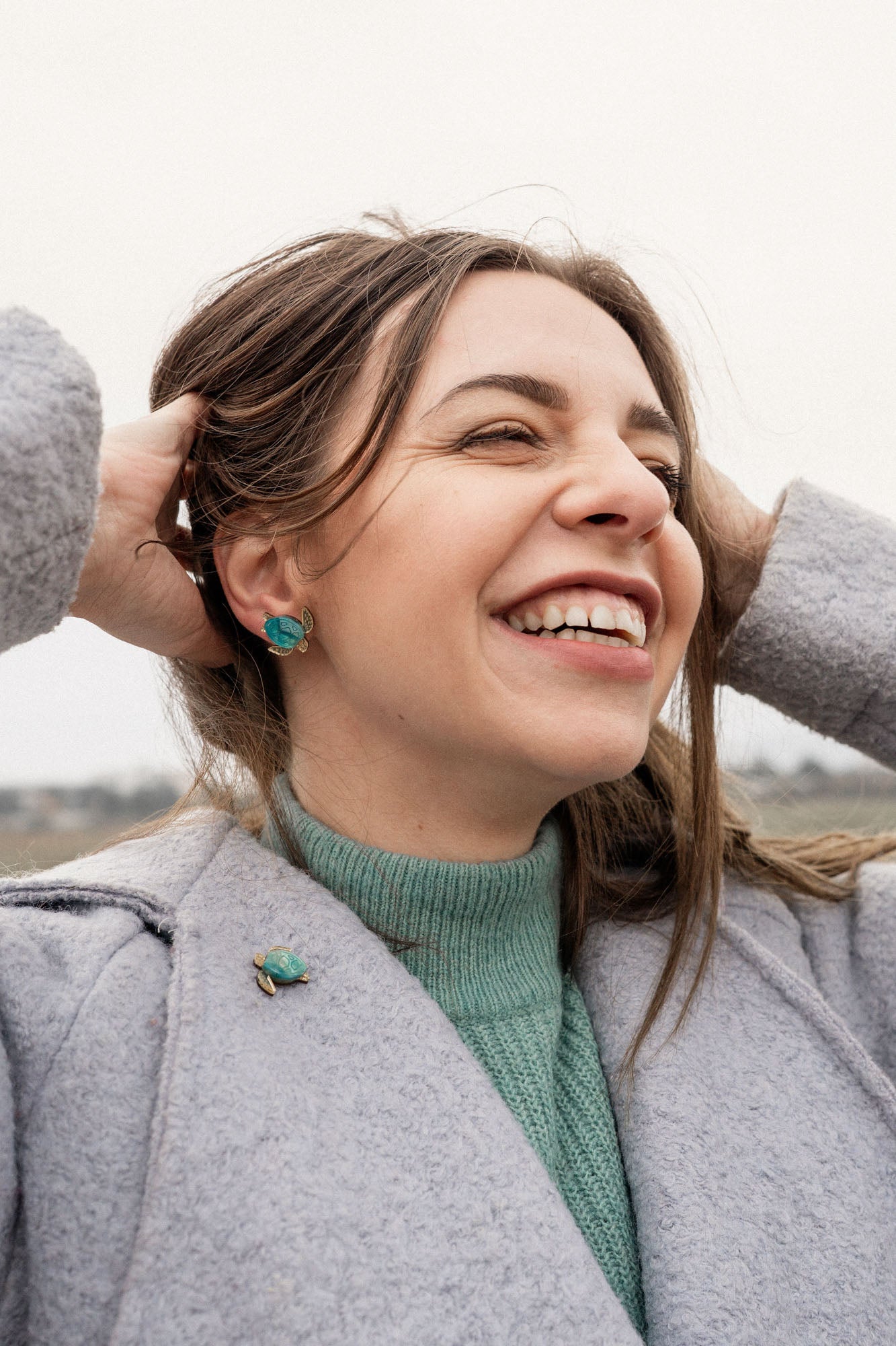 A woman with straight, light brown hair and light skin wears a light pink top and turquoise Sea Turtle Studs, standing in front of a mauve curtain, gazing slightly to the left for a summery vibe.