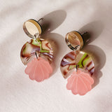 A pair of handmade earrings with round, marbled discs in cream, brown, and green, each featuring a pink, translucent, flower-shaped charm hanging below; lightweight and displayed on a light textured surface with soft shadows.