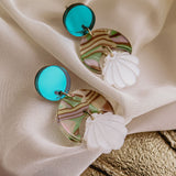 A pair of handmade Seashell Earrings featuring round turquoise studs, marbled circular pieces in green, pink, and clear tones, and lightweight white seashell-shaped charms, displayed on draped white fabric.