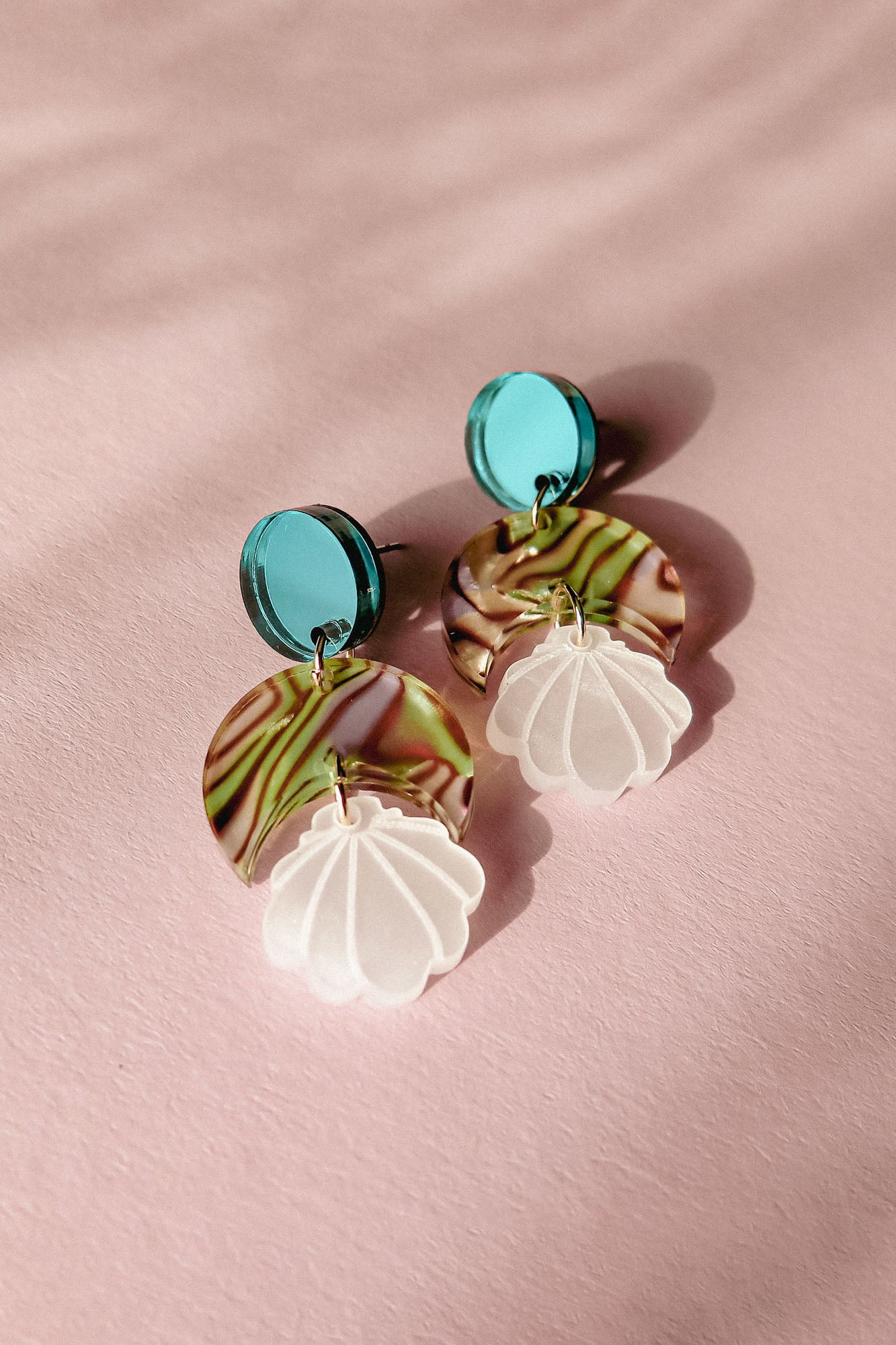 A pair of handmade Seashell Earrings with turquoise circular studs, marbled green and brown semicircle pieces, and translucent white shell-shaped danglers, displayed on a pink textured surface. Lightweight and perfect for any occasion.