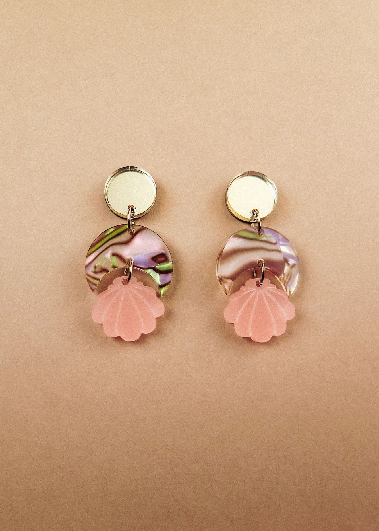 Handmade gold and marbled pink Seashell Earrings with circular tops, lightweight translucent marbled middles, and pink shell-shaped pendants, displayed on a light beige background.